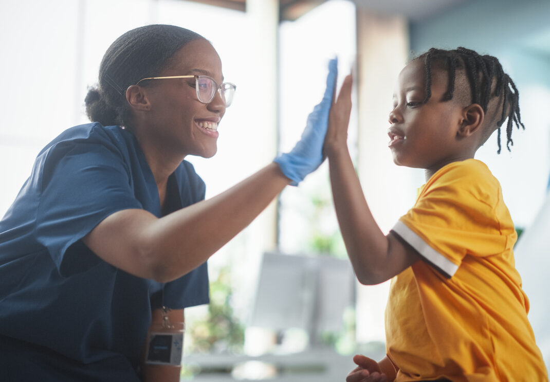 Young African American Boy Sitting In The Chair In Bright Hospital And Getting His Flu Vaccine. Female Black Nurse Is Finished Performing Injection. Professional Woman High-Fives A Kid For Being Brave